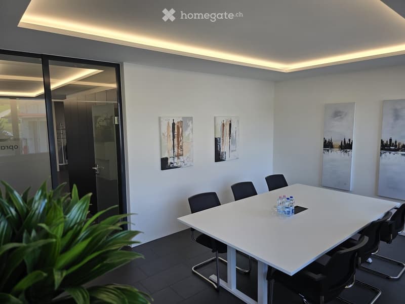 Modern conference room with white table, black chairs, and several water bottles on the table. Artwork is displayed on the walls, and a large glass door leads to another area.