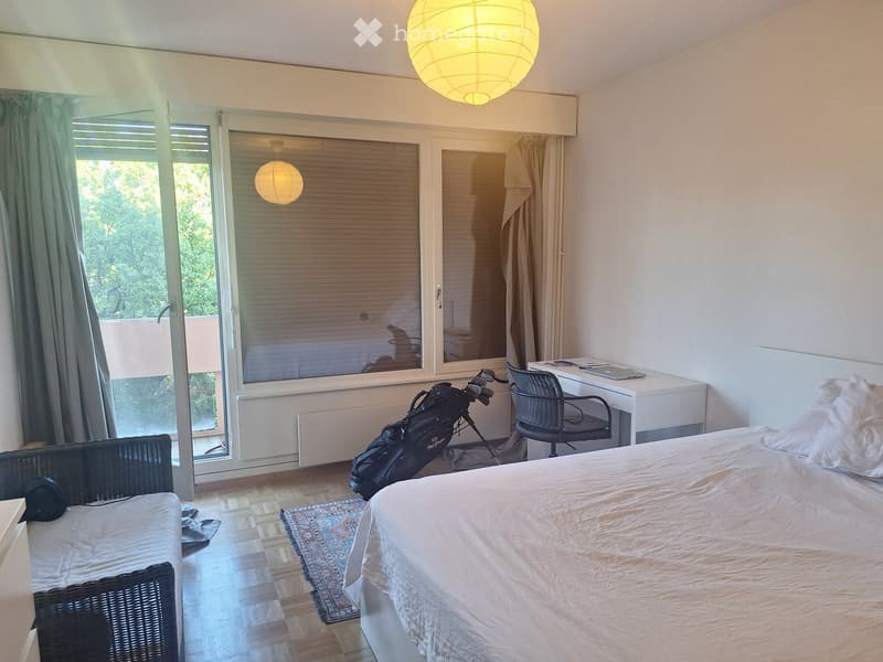 Bright bedroom with a large bed, wicker sofa, desk, and golf bag near the window. The room features wooden flooring, a patterned rug, and large windows with blinds.