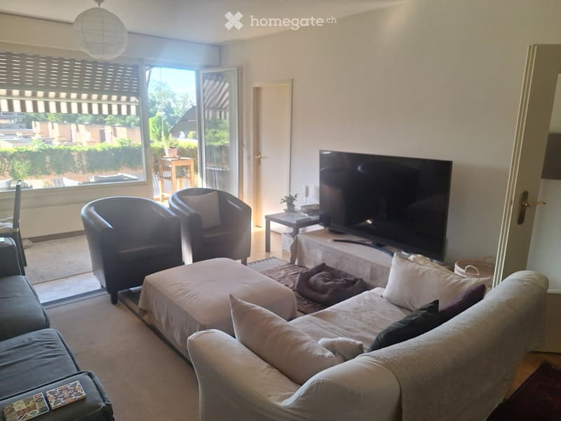 Bright living room with large sliding glass doors leading to a balcony, a grey sectional sofa with an ottoman, two armchairs, a coffee table, a television on a stand, and a side table with a lamp.