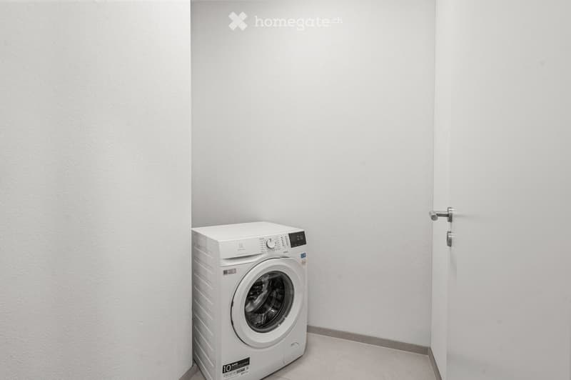 A small, empty room with white walls and a tiled floor, featuring a single white washing machine placed against the wall.