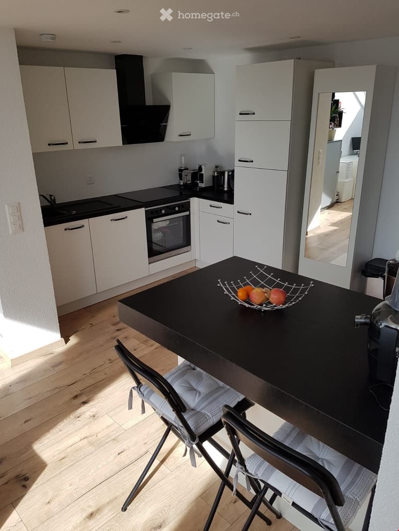 Modern kitchen with white cabinets, black countertops, and stainless steel appliances. Includes a coffee maker, fruit bowl, and a small dining area with a table and chairs.