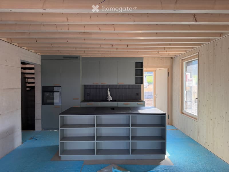 Modern kitchen with grey cabinets, black countertop, built-in oven, and open shelving unit. Exposed wooden beams on the ceiling and large windows provide natural light.