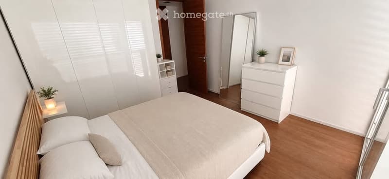 Modern bedroom with a large bed, white nightstand, mirror, dresser, and small potted plants.