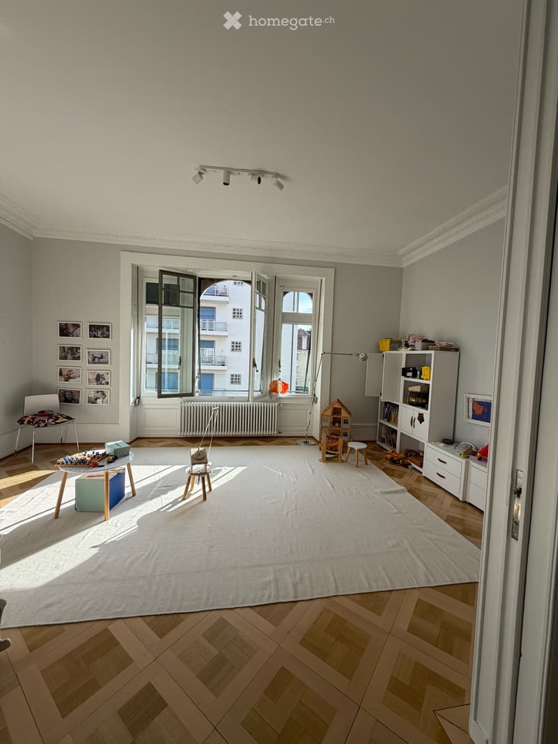 Bright, airy room with wooden herringbone flooring, large white rug, large windows with open shutters, and various children's furniture and toys.