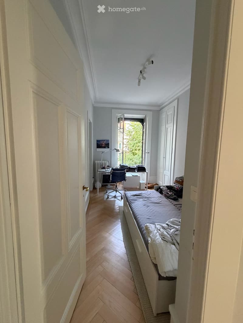 Bedroom with wooden herringbone flooring, bed with grey duvet, desk with chair, radiator, and a screen on the wall.