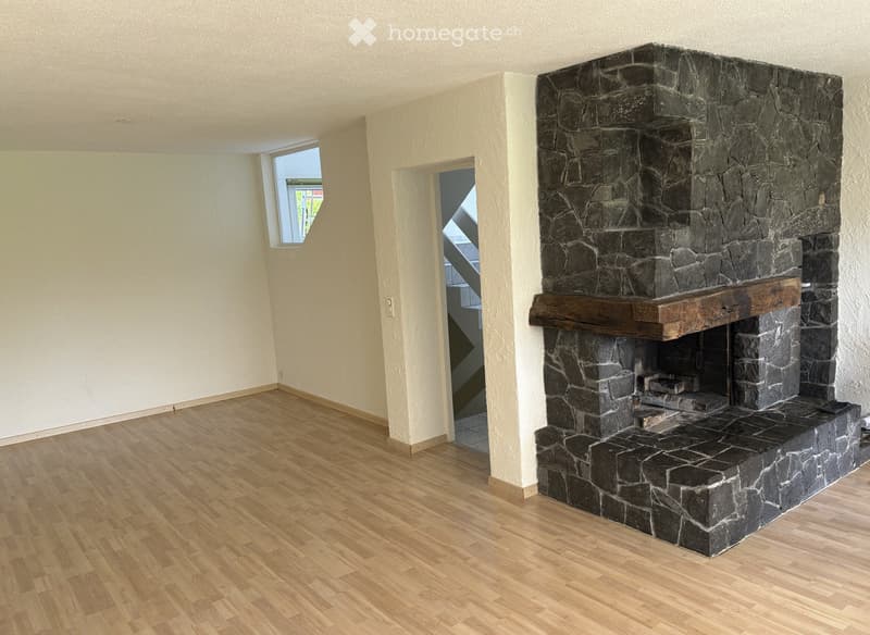 Empty room with wooden flooring, white walls, and a stone fireplace with a wooden mantel. A small window and an open doorway lead to another area.