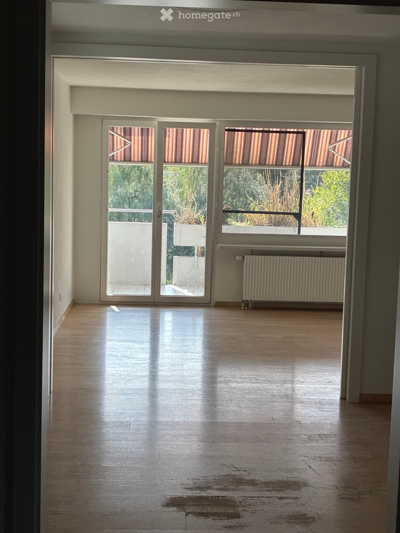 Empty room with wooden flooring, large sliding glass doors leading to a balcony, radiator, and visible outdoor greenery.