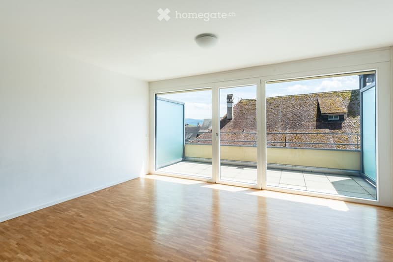 Empty room with wooden flooring, white walls, and large glass sliding doors leading to a balcony with a view of rooftops.