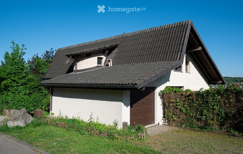 Two-story house with a dark tiled roof, white walls, and a wooden garage door. The house is surrounded by greenery and has a small garden area.