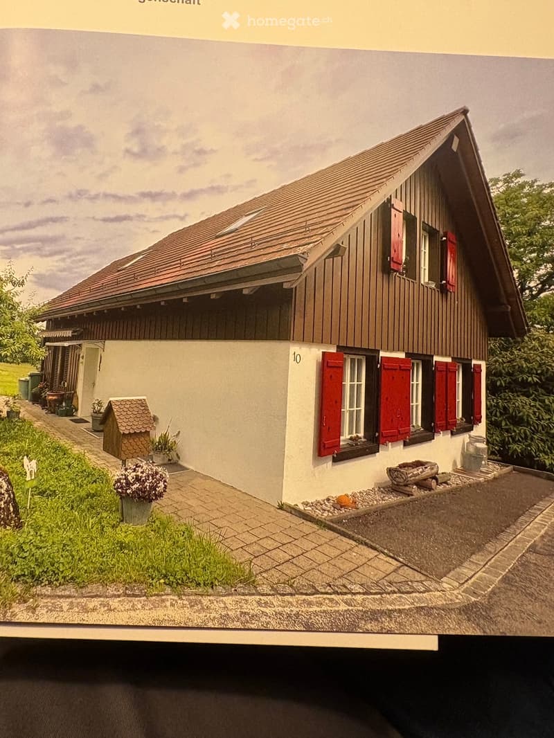 Two-story house with a red-tiled roof, white walls, and red shutters. The house features a small balcony and is surrounded by a garden with potted plants and a small doghouse.