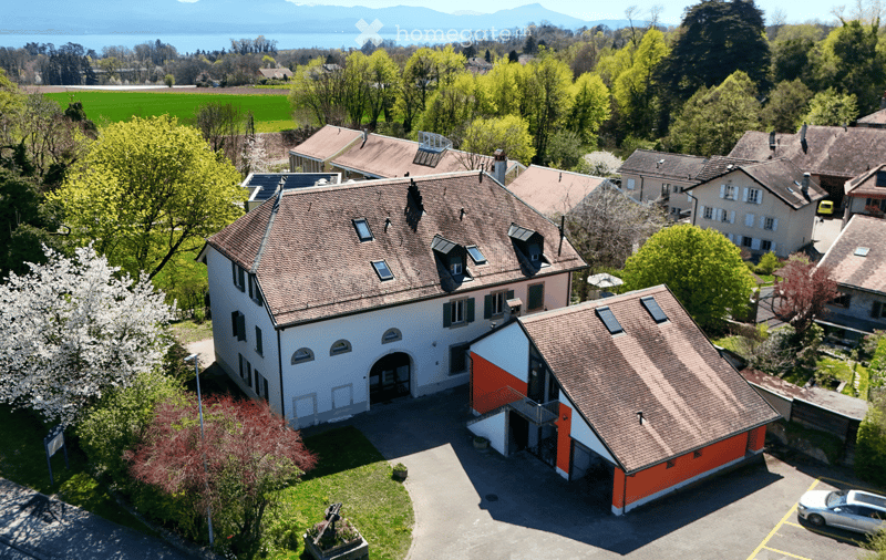 Aerial view of a large residential building with a garage and parking area, surrounded by greenery and neighboring houses.