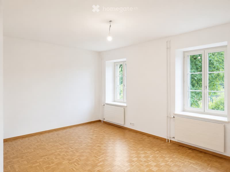 Empty room with light-colored walls, wooden herringbone flooring, and two windows with white frames. Radiators are present beneath each window.