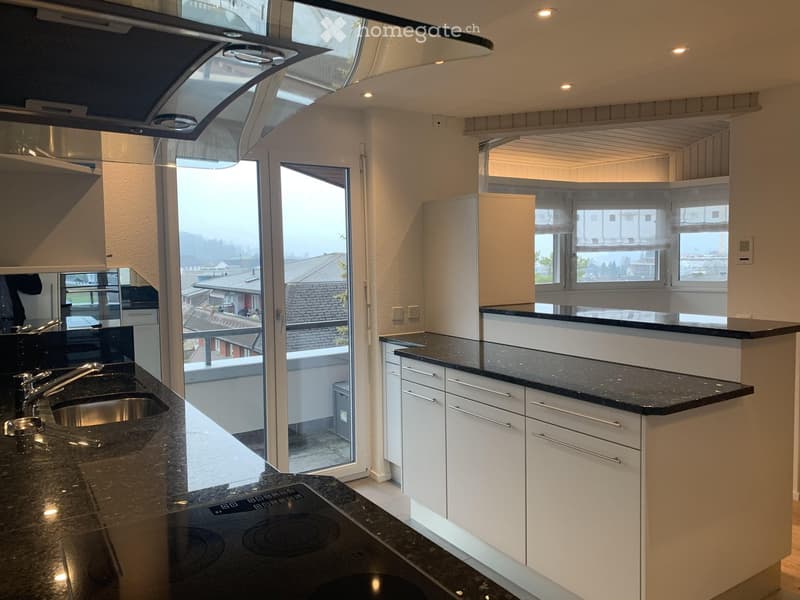 Modern kitchen with white cabinets, black countertops, stainless steel sink, and built-in stove. Large windows and sliding glass door lead to a balcony.