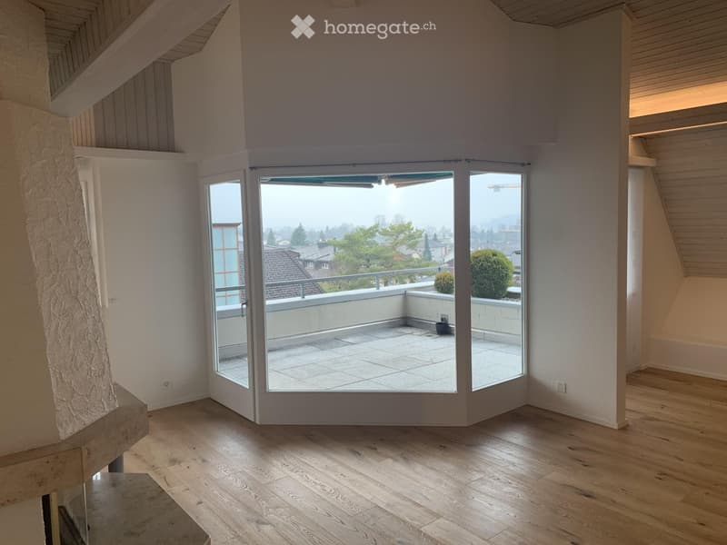 Bright, empty room with wooden flooring, large sliding glass doors leading to a tiled balcony, and a view of the city and surrounding greenery.