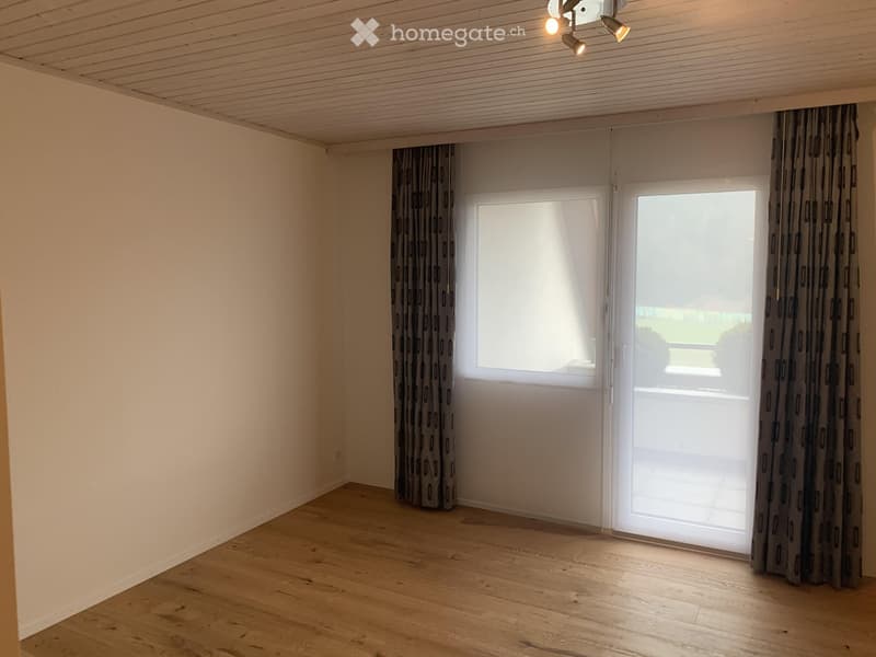 Empty room with wooden flooring, white walls, large window with curtains, and a sliding glass door leading outside.