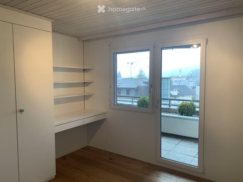 Empty room with wooden flooring, white walls, built-in shelves, and large windows offering a view of the city and a small balcony area.