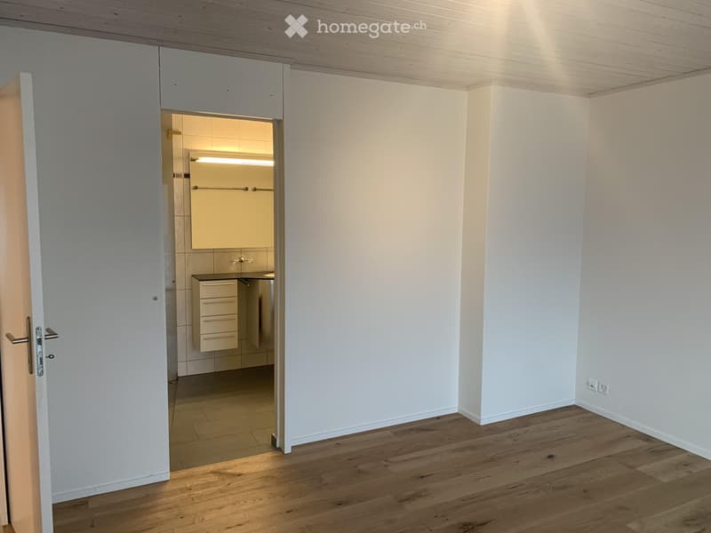 Empty room with wooden flooring and white walls, featuring an open doorway leading to a small, modern bathroom with white cabinets and a sink.