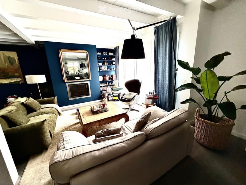 Living room with a beige sofa, coffee table, armchair, floor lamp, and a large plant in a basket. The room features a fireplace, a bookshelf, and a modern design with dark blue walls.