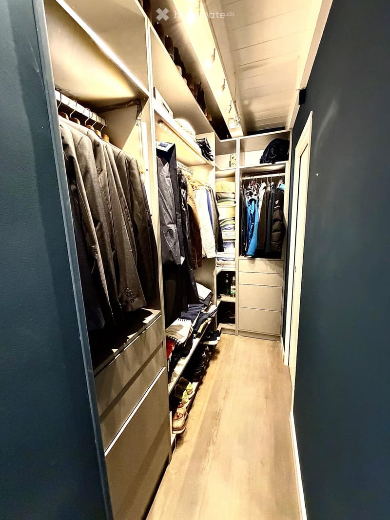 Well-lit hallway with built-in closet units on one side, containing hanging clothes, shelves, and storage compartments. The floor is light-colored wood, and the walls are painted dark blue.