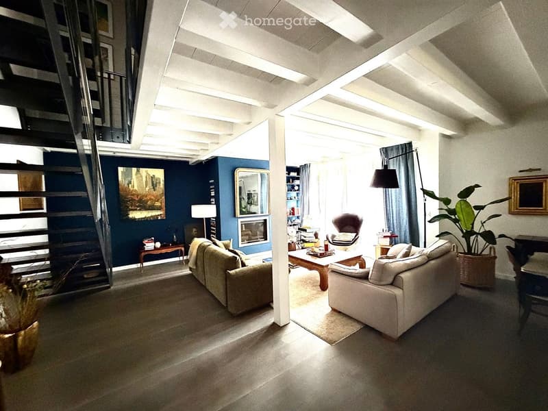 Modern living room with exposed wooden beams, large windows, and a mix of seating including a sofa, armchair, and coffee table. Features a fireplace, bookshelf, and decorative plants.