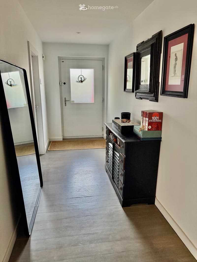 Hallway with wooden flooring, white walls, a black cabinet with drawers, and several framed mirrors and artwork on the wall. A door leads to another room, and a small rug is placed near the entrance.
