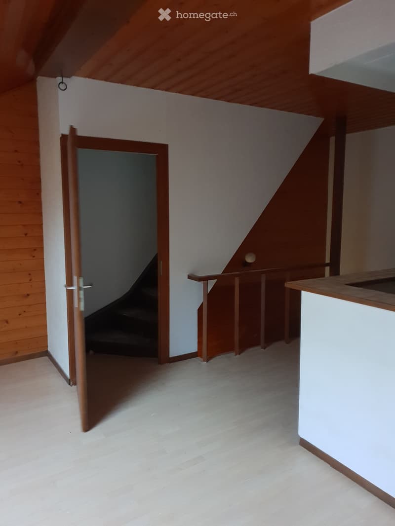 Empty room with wooden floor, white walls, and a wooden door leading to a staircase. The ceiling is slanted with wooden beams.