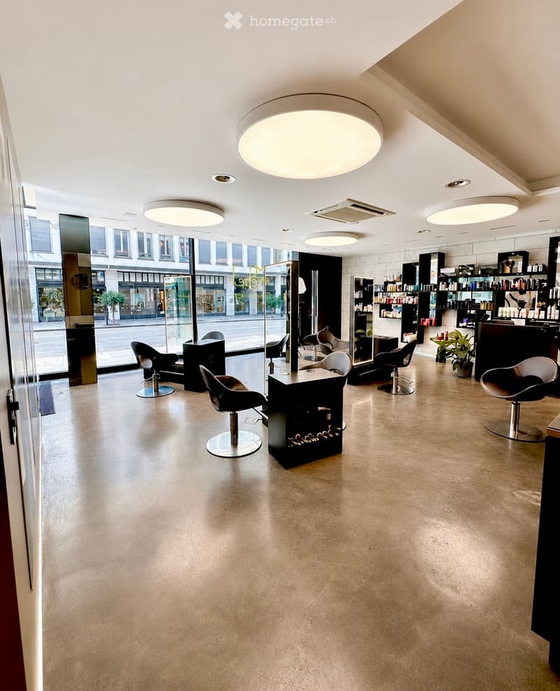 Modern, well-lit salon with polished concrete floors, multiple stylist chairs, a central mirror, shelves with hair products, and a reception desk.