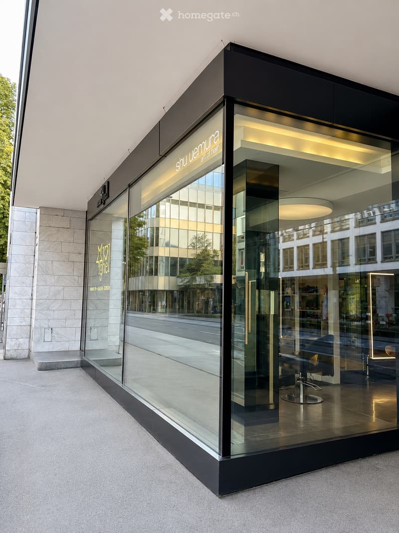 Modern storefront with large glass windows and black framing, displaying the name "Shu Uemura" and interior lighting. The reflection shows a street and surrounding buildings.