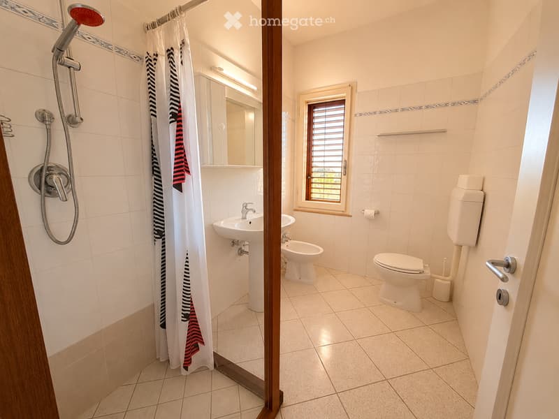 Modern bathroom with white tiles, a toilet, a sink, a shower with a curtain, and a window with blinds.