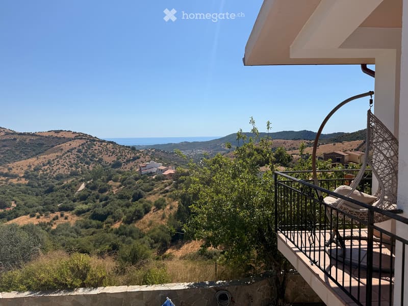 Balcony with a wicker hanging chair, overlooking a scenic landscape with hills, trees, and the sea.