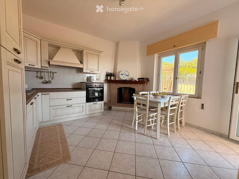 Bright kitchen with white cabinets, a central island, a built-in oven, a fireplace, and a dining area with a table and chairs. The space features tiled flooring and large windows offering a view of the outdoors.
