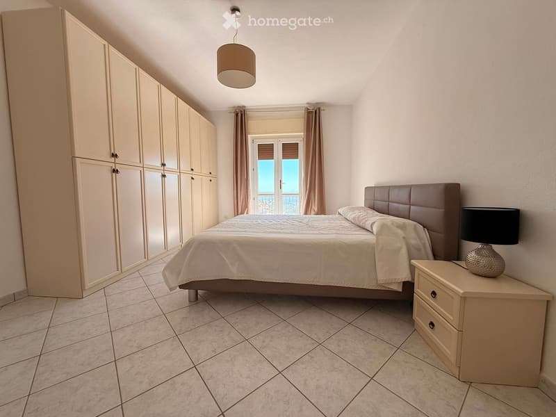 Modern bedroom with a large bed, nightstand, lamp, and built-in wardrobe. The room features tiled flooring and large windows with curtains.