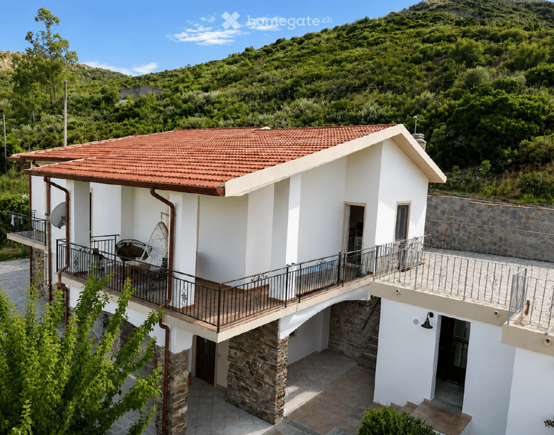 Two-story white house with red-tiled roof, stone accents, and balconies overlooking a green, hilly landscape.