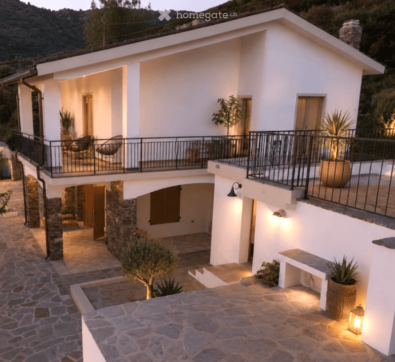 Two-story white house with a balcony, stone base, and outdoor seating area.