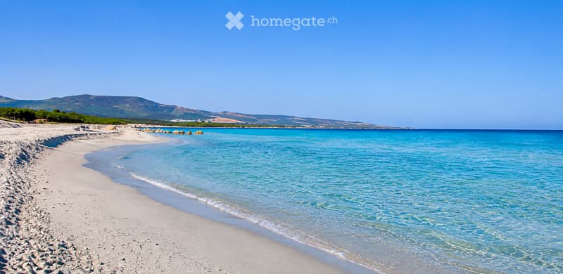 A serene beach with fine white sand, clear turquoise water, and distant hills under a bright blue sky.