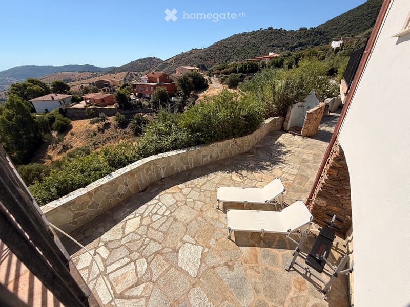 Balcony with stone flooring, two sun loungers, and a fitness machine, overlooking a scenic hillside with houses and greenery.