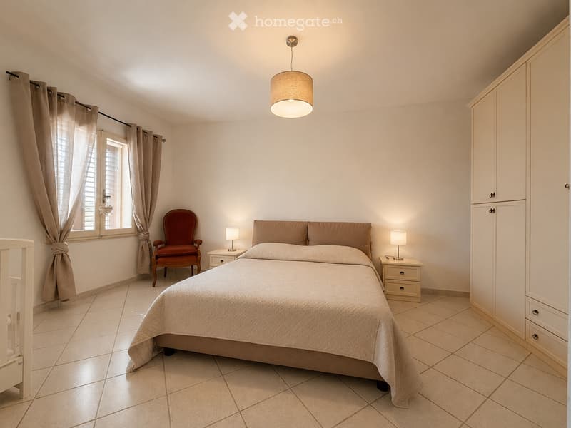 Cozy bedroom with a double bed, beige bedding, a wooden wardrobe, a red armchair, and bedside tables with lamps.