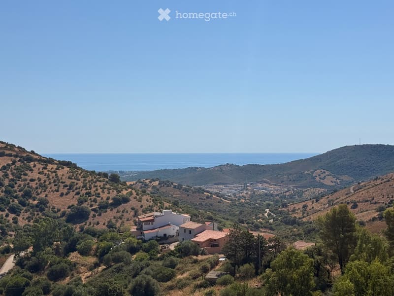 A panoramic view of a residential area nestled in a hilly landscape, with several houses and a clear view of the sea in the distance.