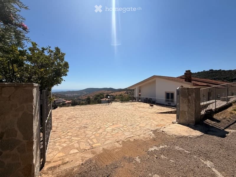 A spacious outdoor area with a stone-paved courtyard, a white house in the background, and a scenic mountain view under a clear blue sky.