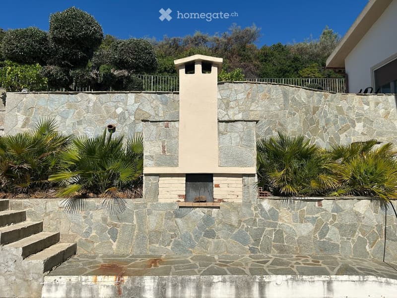 Stone wall with a small chimney structure, surrounded by palm trees and greenery, with steps leading up to the wall.