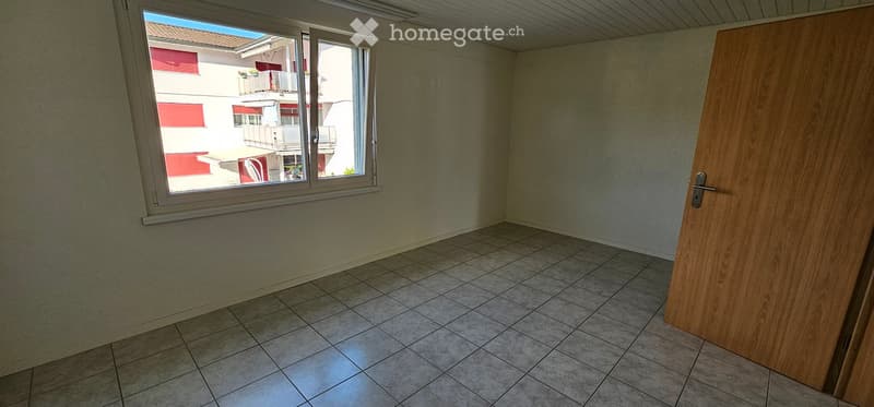 Empty room with tiled floor, white walls, and a large window offering a view of a neighboring building with balconies.