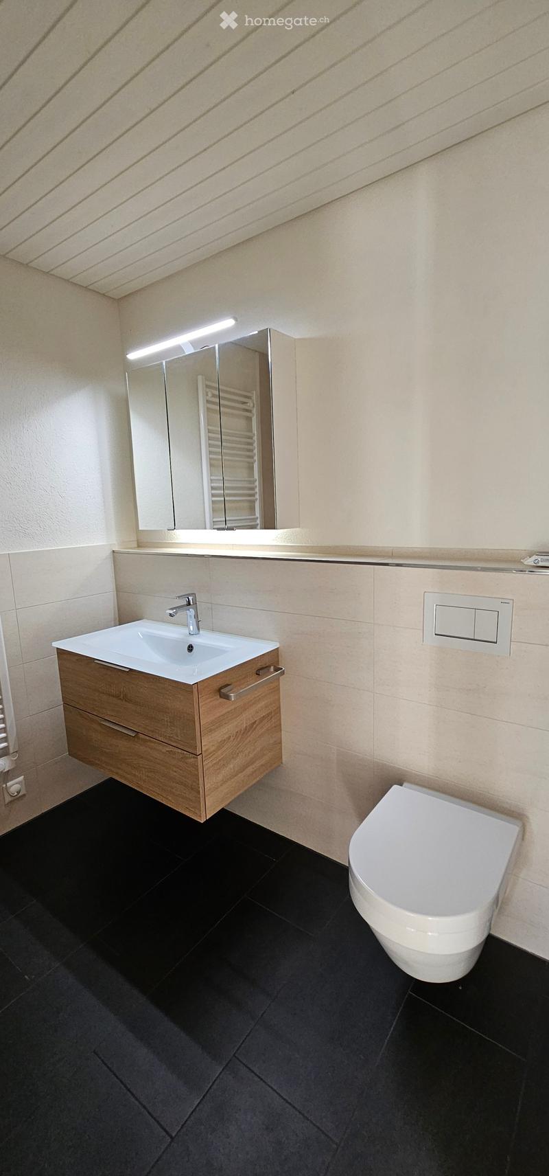 Modern bathroom with a white sink, wooden cabinet, chrome faucet, large mirror with built-in lighting, and a white toilet. Black tiled floor and beige tiled walls.