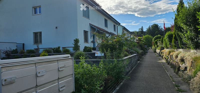 A narrow pathway lined with greenery leads to a residential building with light blue walls and multiple windows. The area is well-maintained with various plants and shrubs, and a flag is visible on a pole in the distance.