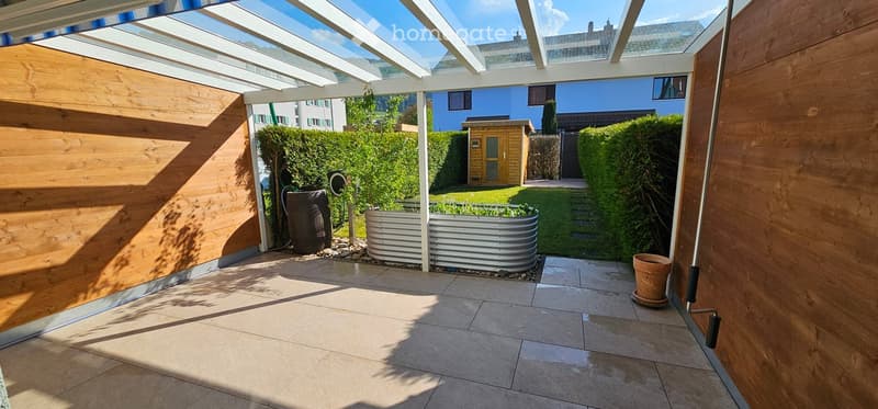 A covered patio area with tiled flooring, wooden walls, and a metal planter in the center. The background shows a well-maintained garden with hedges, a small wooden shed, and a blue house.