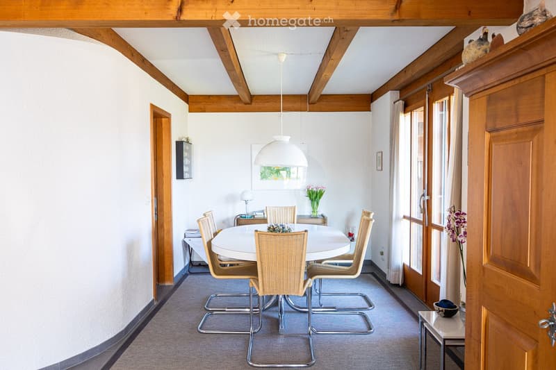 Dining room with round white table, wicker chairs, wooden beams, white walls, glass doors, flowers, lamp