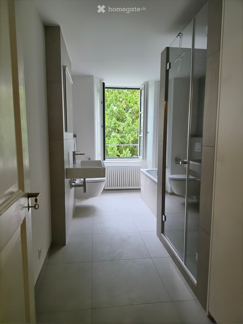 Modern bathroom with white tiles, a sink, toilet, bathtub, and a large window offering a view of greenery.