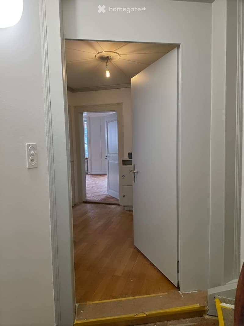 A narrow hallway with wooden flooring, white walls, and a ceiling light. An open door leads to another room with visible furniture and a tiled floor.