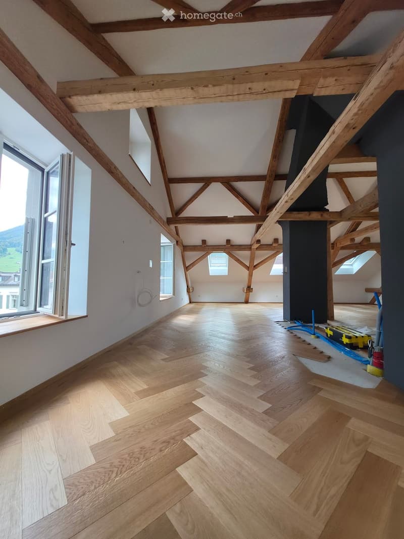 Empty attic space with exposed wooden beams, wooden flooring, and large windows allowing natural light. No furniture or equipment present.