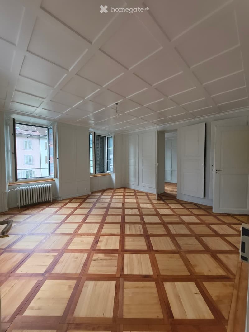 Empty room with wooden parquet flooring, white walls, large windows, and a radiator. The ceiling features a decorative, coffered design with integrated lighting.