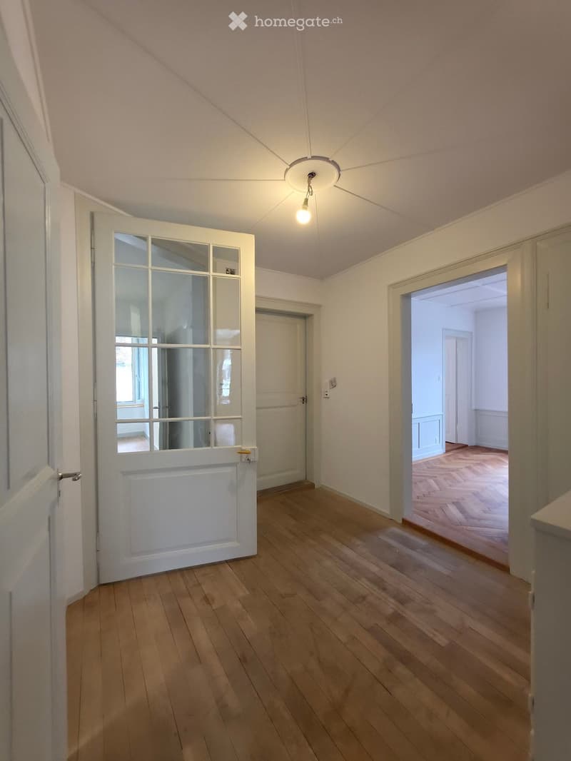 Empty hallway with wooden flooring, white walls, and a ceiling light fixture. Doors lead to adjacent rooms.